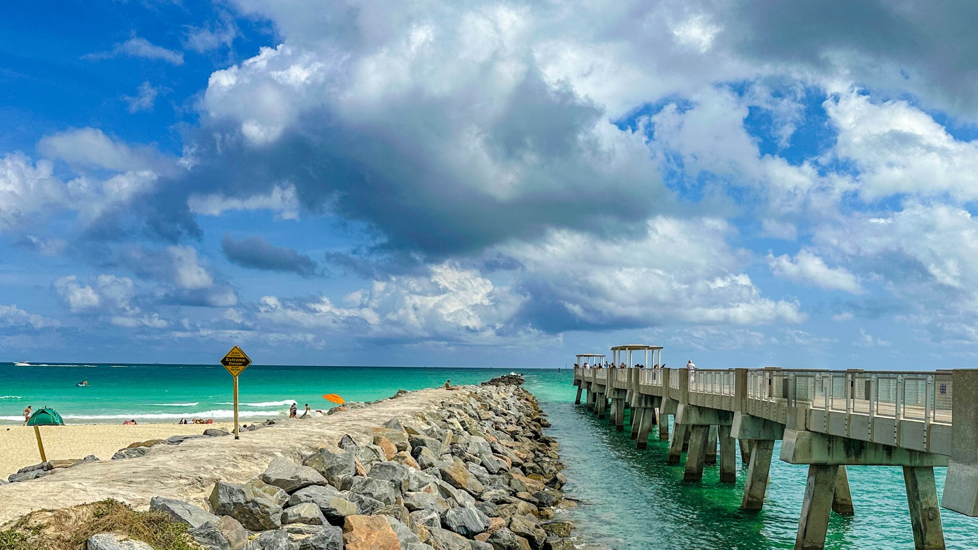 Rock jetty beside a fishing pier on a sunny beach with turquoise water and a warning sign