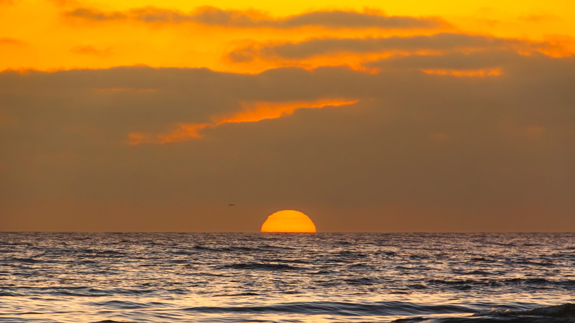 Orange sun setting into the ocean horizon beneath a cloudy golden sky