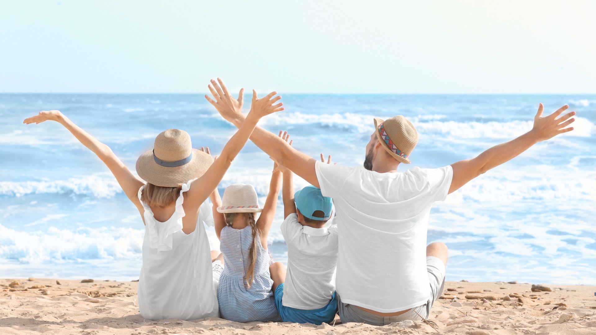 Family of four sitting on the sand with arms raised, facing ocean waves on a sunny day