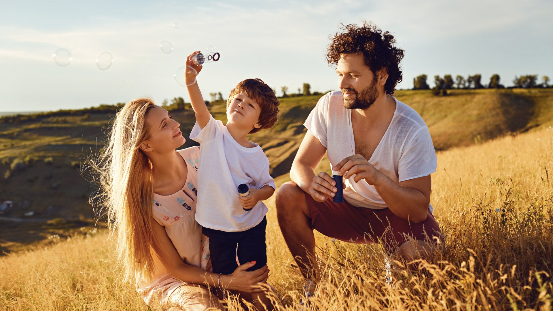 Mother, father, and young son blowing bubbles together in a golden grassy field at sunset
