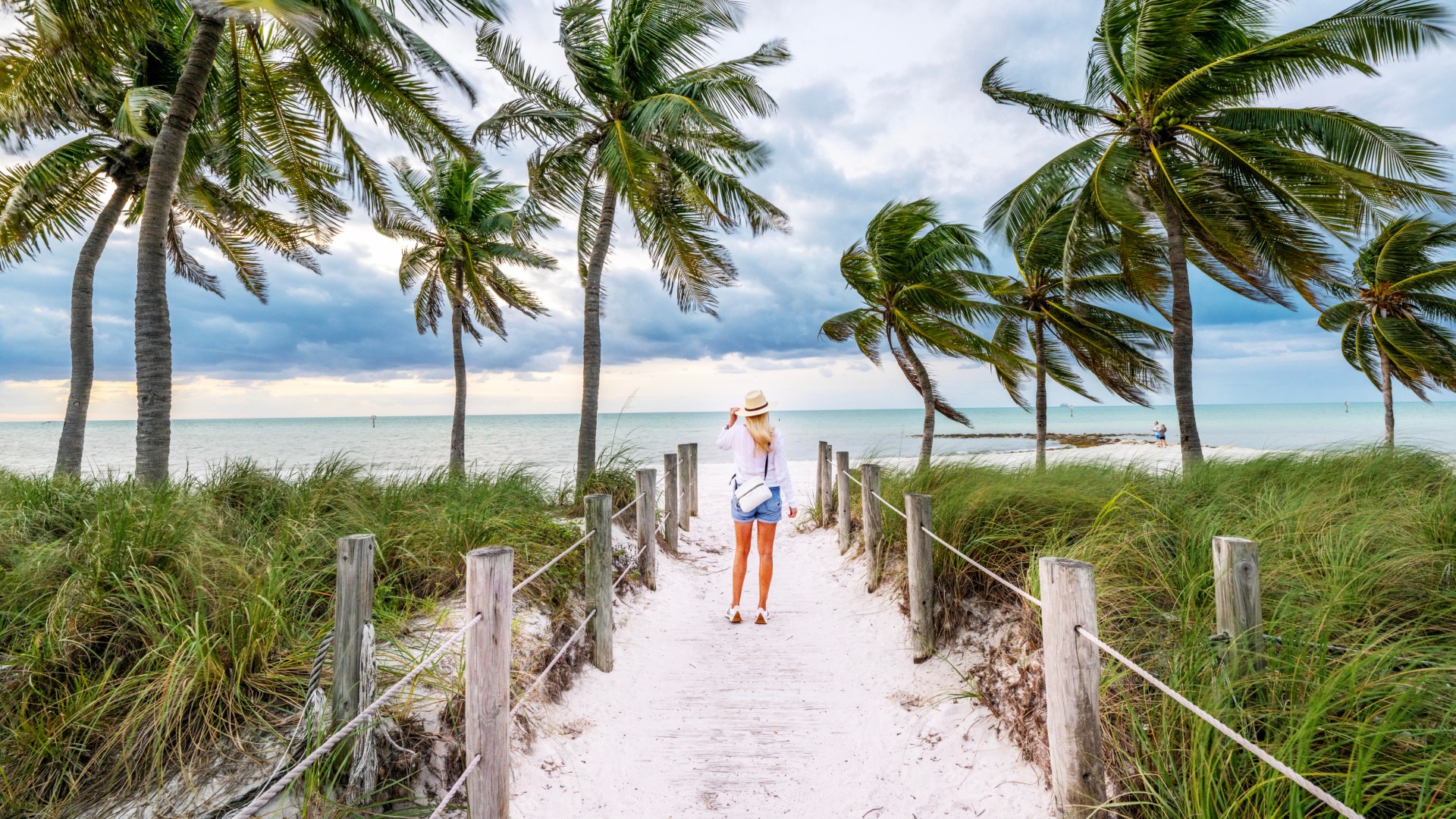 Woman in a sun hat walking a sandy beach path lined with rope fencing and windblown palm trees