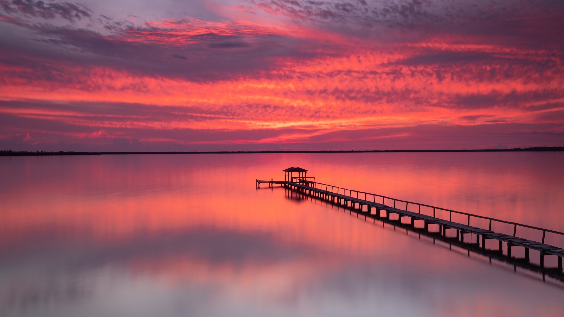 Wooden pier stretching into calm water under a vivid pink and red sunset near West Palm Beach, Florida