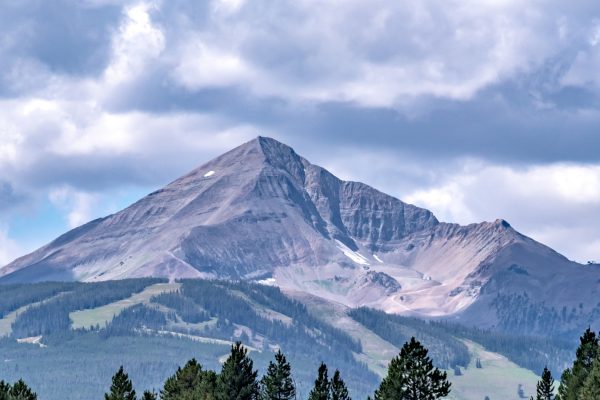 Snow-dusted mountain peak rising above evergreen forests under dramatic cloudy skies.