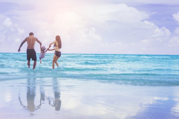 Parents holding their child’s hands while playing together in shallow ocean water at the beach.