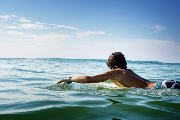 Person paddling on a surfboard in calm ocean water under a bright blue sky.