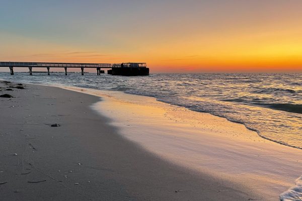 Sunset over a sandy beach with gentle waves and a long pier extending into the ocean.
