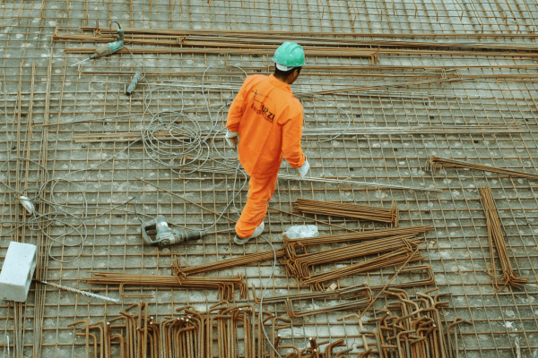 obrero de la construcción vestido con un mono naranja brillante y un casco verde, caminando por una cimentación de rejilla metálica, que está cubierta de barras de refuerzo y herramientas de construcción.