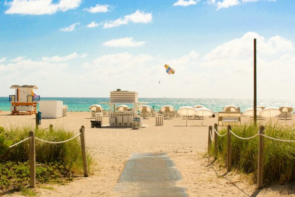 Sandy beach entrance leading to umbrellas, lifeguard towers, and turquoise ocean under a sunny sky.