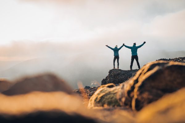 Two people standing on a rocky mountain overlook with arms raised.