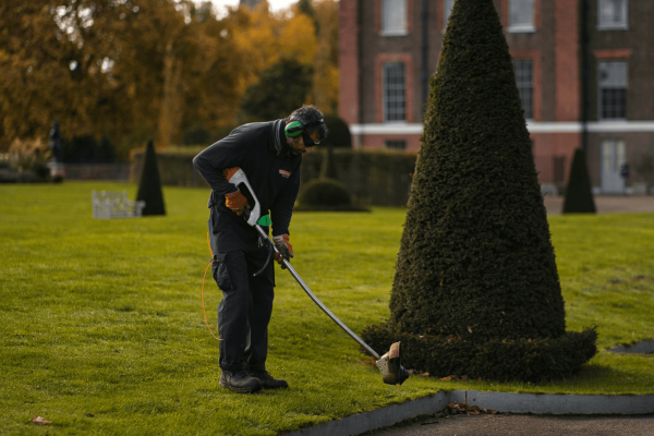 A man in a navy jumpsuit using a weed eater/lawn trimmer on a lawn