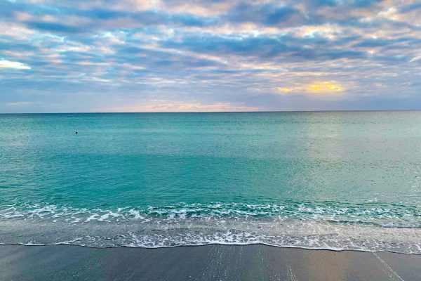 Calm turquoise ocean water meeting a sandy beach beneath a cloudy sunrise sky.