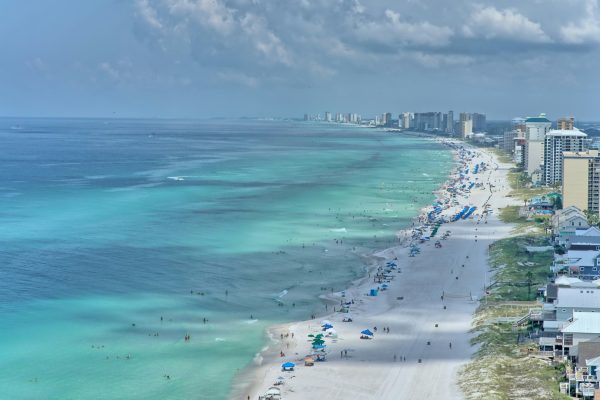 Aerial view of a crowded beachfront with turquoise ocean water, white sand, and high-rise buildings along the coast.