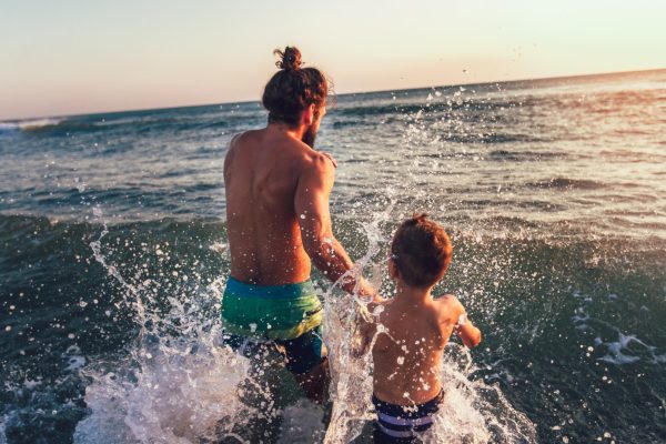 Father and son running into ocean waves together at sunset with water splashing around them.