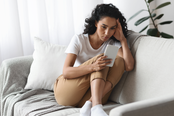 A young woman with curly hair sitting on a couch, looking at her phone with a concerned expression, resting her head on her hand in a moment of distress.