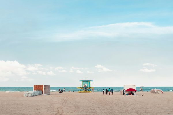 Wide sandy beach with a colorful lifeguard tower, a few beachgoers, and the ocean beneath a bright blue sky with soft clouds.