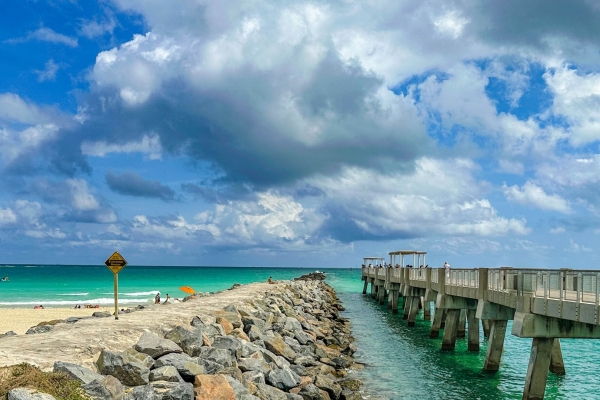 Un espigón de rocas junto a un muelle de pescadores en una playa soleada con aguas turquesas y una señal de advertencia