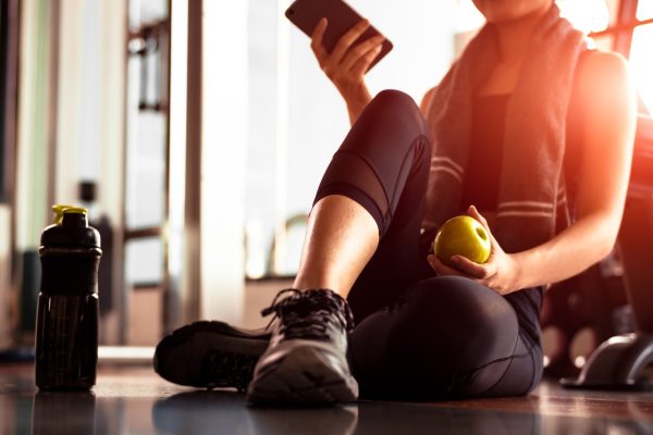 Person sitting on a gym floor holding a phone and an apple next to a water bottle.