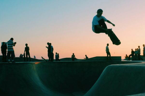Skateboarder performing a high jump trick at a skatepark during sunset with other skaters in the background