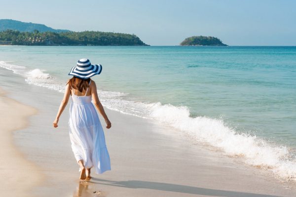 Woman in a white dress and wide-brim hat walking barefoot along a tropical beach shoreline.