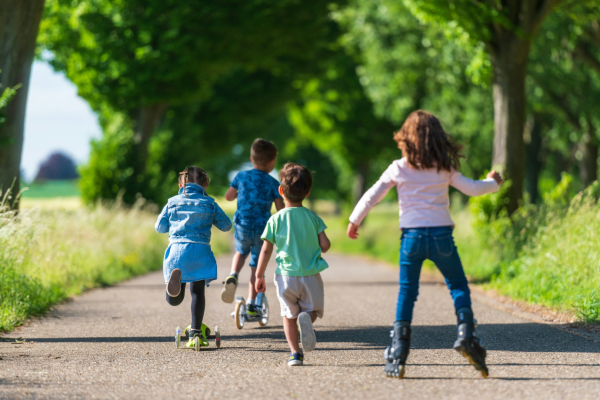 A group of children playing outdoors on a tree-lined pathway, riding scooters, rollerblading, and running, enjoying a sunny day in nature.