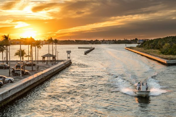 Boat speeding through a coastal inlet at sunset with palm trees, docks, and shoreline in the background.