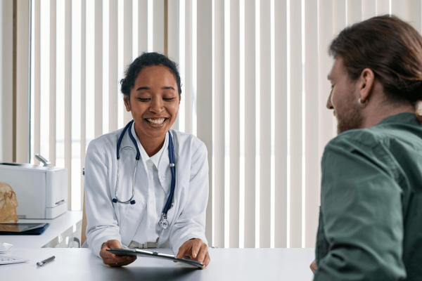 Un amable médico, con bata blanca y estetoscopio, sonríe sentado frente a un paciente en un despacho. El médico sostiene una tableta, probablemente revisando o compartiendo información médica con el paciente, que está sentado de espaldas a la cámara. El paciente lleva el pelo largo recogido en un moño y viste una camisa verde. El ambiente es de colaboración y tranquilidad.