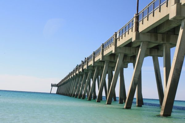 Long concrete fishing pier extending over calm turquoise ocean water beneath a clear blue sky.