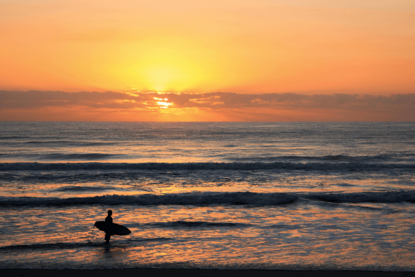 A photo of a warm sunset over the ocean. In the bottom left hand corner is a surfer holding a surfboard looing out at the horizon