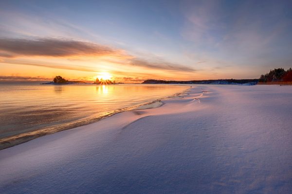 shoreline at sunset with calm water reflecting warm light and a distant tree-lined coast.
