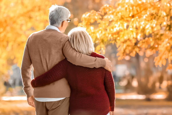 An elderly couple walking together in a park during autumn, with the man’s arm around the woman’s shoulder, showing affection and support.