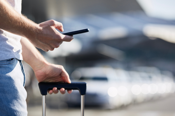 A man holding a suitcase and looking at his phone while waiting for a rideshare in an airport parking area.