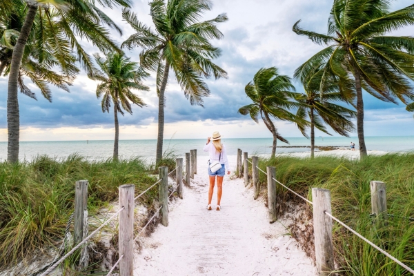 Una mujer con un sombrero para el sol que camina por un sendero de arena en la playa, bordeado por una valla de cuerda y palmeras mecidas por el viento
