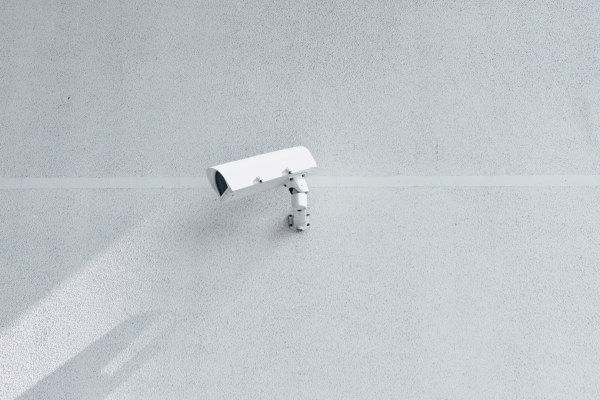 A white surveillance camera mounted on a plain white wall, capturing its surroundings.