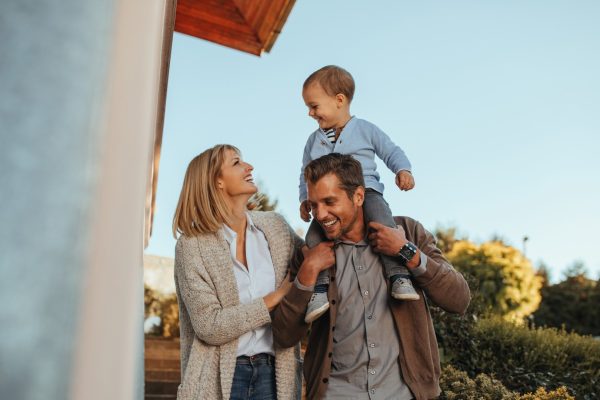Happy parents walking outside with their young child sitting on the father’s shoulders, smiling together.