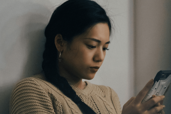 Foto de una mujer joven con el pelo oscuro trenzado sobre el hombro, mirando atentamente su teléfono. Lleva un jersey de punto beige y unos sencillos pendientes de aro. Su expresión es tranquila y concentrada mientras interactúa con su dispositivo, sentada contra una pared de color neutro. La suave iluminación crea un ambiente pensativo y reflexivo.