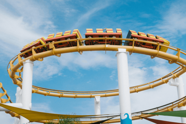 A brightly colored roller coaster, with red, yellow, and orange cars, traveling along a curving yellow track. The track is supported by tall white columns, and the ride takes place under a clear blue sky with light clouds. There are colorful canopies visible at the bottom of the image, possibly part of a theme park or fairground setting. The perspective looks up toward the roller coaster, capturing the motion and excitement of the ride.