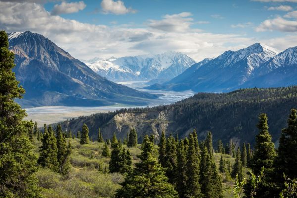 Scenic mountain valley with snow-capped peaks, evergreen forest, and a winding river under a partly cloudy sky.