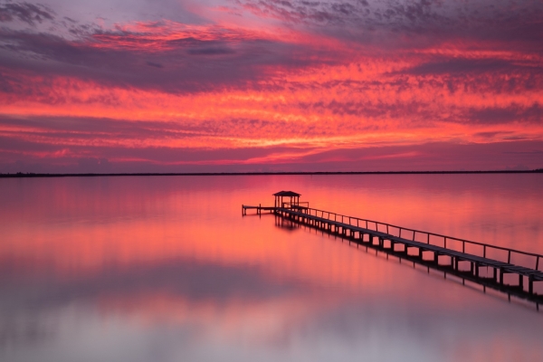 Un embarcadero de madera que se adentra en aguas tranquilas bajo una puesta de sol de intensos tonos rosas y rojos, cerca de West Palm Beach, Florida