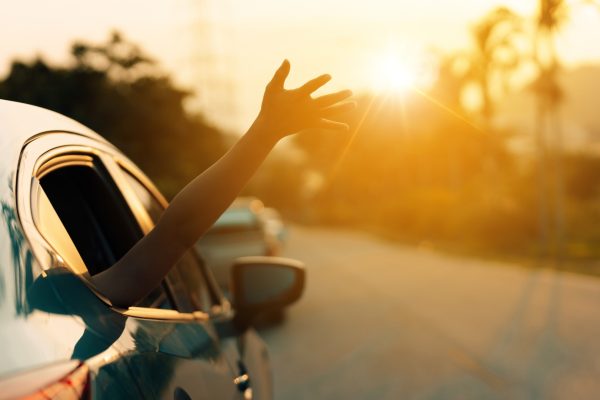 Person’s arm extended out of a car window toward the sun while driving down a road at sunset.