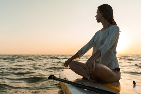 Woman sitting cross-legged on a paddleboard in the ocean at sunset, looking out over the water.