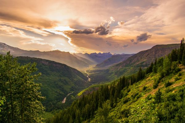 Scenic mountain valley at sunset with evergreen forests, distant peaks, and golden light illuminating the landscape.