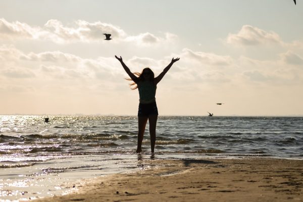 Silhouette of a woman standing at the shoreline with arms raised as seabirds fly overhead during a glowing sunset.