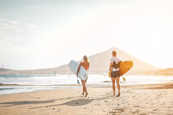 Two surfers carrying surfboards walk along a sunlit beach toward the ocean as the sun sets behind distant hills