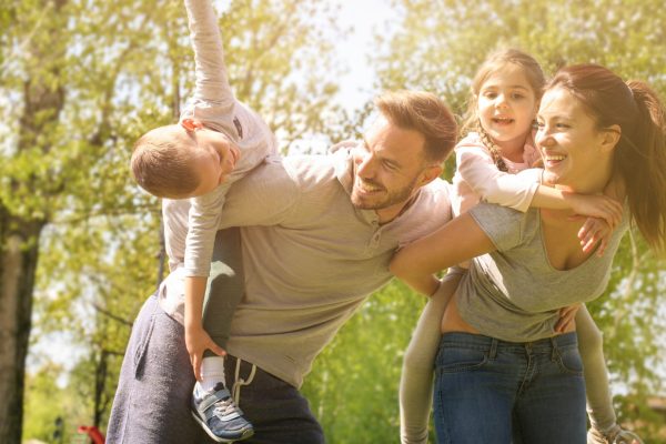 Smiling family of four playing together outdoors as parents give their children piggyback rides in a sunny park.