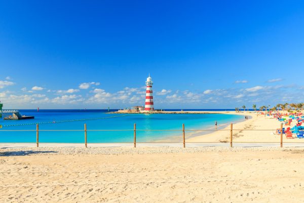 Red and white striped lighthouse overlooking a calm turquoise beach with white sand and clear blue skies.