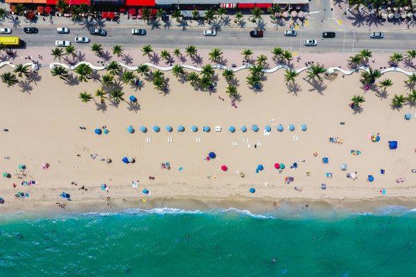 Aerial view of a sandy beach lined with palm trees, colorful umbrellas, and turquoise ocean water beside a coastal roadway.